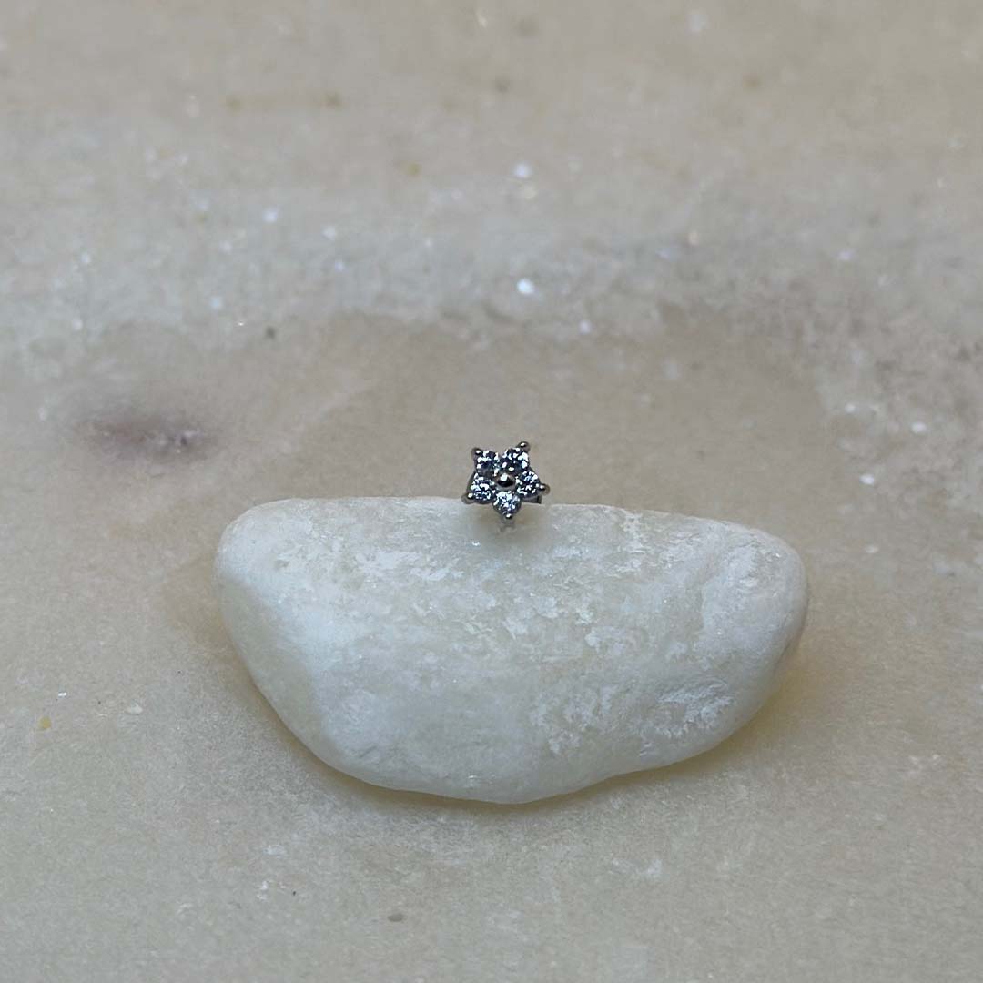 Un très beau piercing "hiny", de couleur argent, est au centre de l'image. Le bijoux en acier inoxydable est posé de face sur un caillou blanc. Le fond est du sable blanc.
