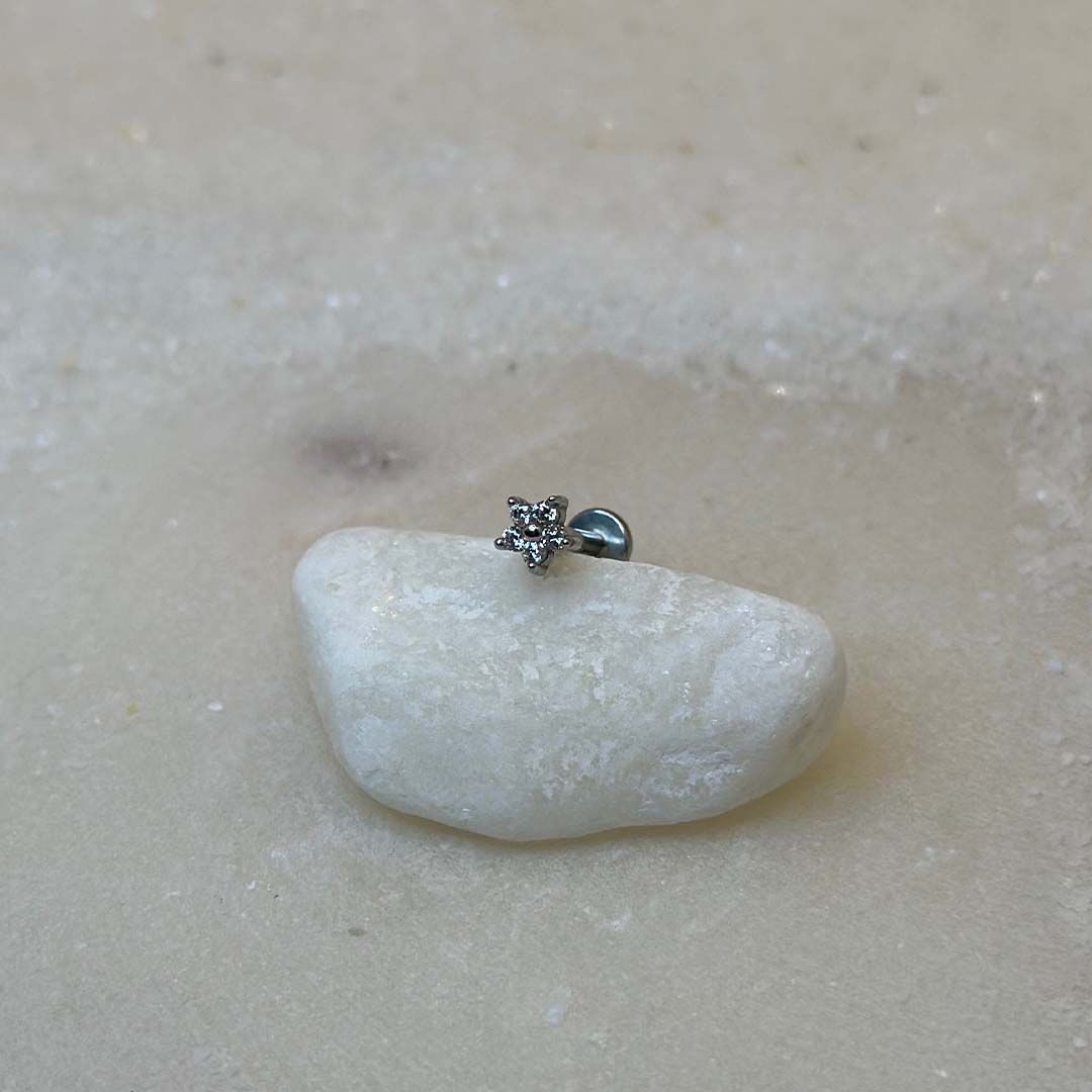 Un très beau piercing "hiny", de couleur argent, est au centre de l'image. Le bijoux en acier inoxydable est posé de 3/4 sur un caillou blanc. Le fond est du sable blanc.