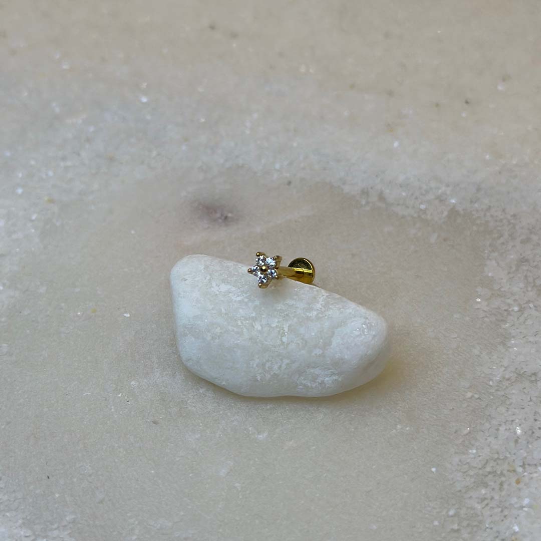 Un très beau piercing "hiny", de couleur or, est au centre de l'image. Le bijoux en acier inoxydable est posé sur un caillou blanc. Le fond est du sable blanc.