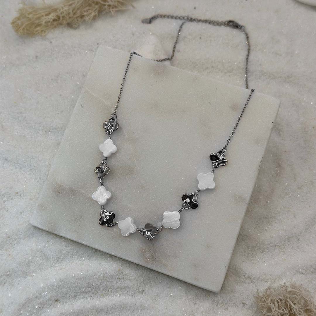 Un jolie collier argenté "Inaya" est au centre de l'image, posé sur une plaque de marbre. Le bijoux en acier inoxydable est composé de trèfles argentés et blancs. Tout autour se trouve du sable blanc et des algues séchées.