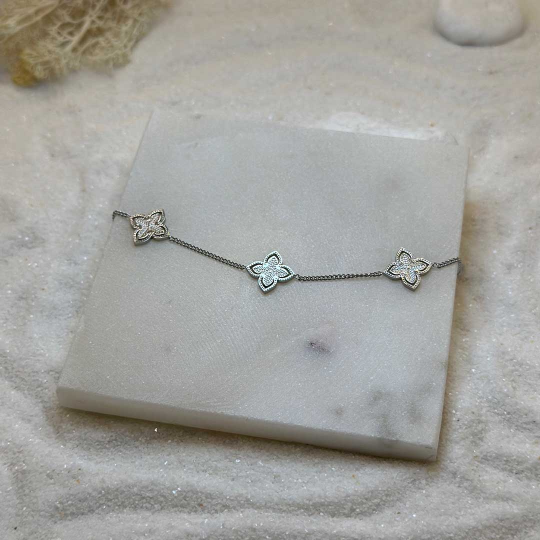Un très jolie bracelet en argent "irina" est au centre de l'image en gros plan, posé sur une plaque de marbre. Le bijoux en acier inoxydable est composé d'une chaîne en argent et deux 3 charmes en forme d'étoiles. Tout autour se trouve du sable blanc.