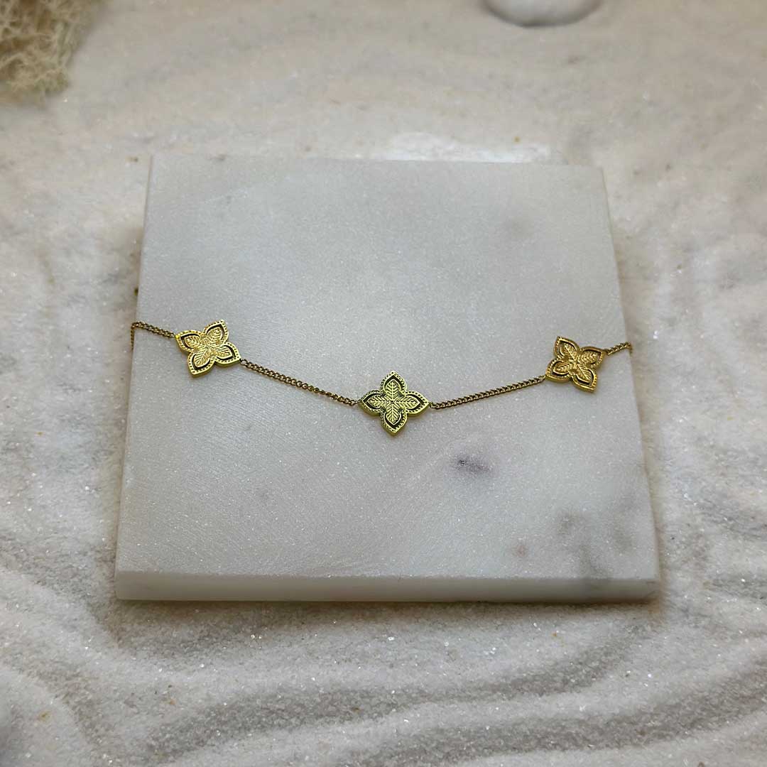 Un très jolie bracelet en or "irina" est au centre de l'image en gros plan, posé sur une plaque de marbre. Le bijoux en acier inoxydable est composé d'une chaîne en argent et deux 3 charms en forme d'étoiles. Tout autour se trouve du sable blanc.