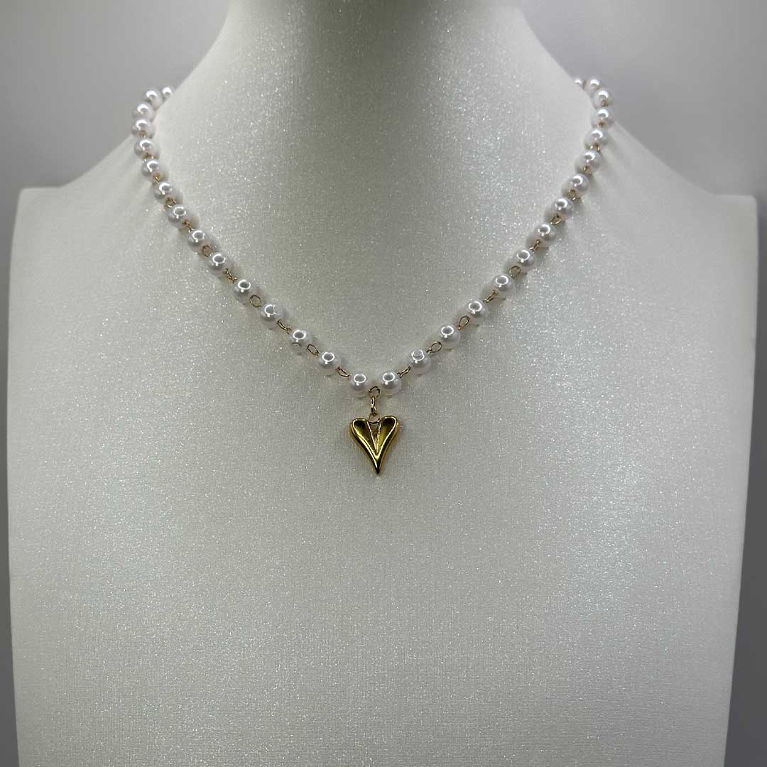 Ce magnifique bijou "Jane" en acier inoxydable est un collier de parles blanches avec un pendentif coeur de couleur or. Il est posé sur un buste pailleté de couleur blanc qui est lui même sur un fond blanc.