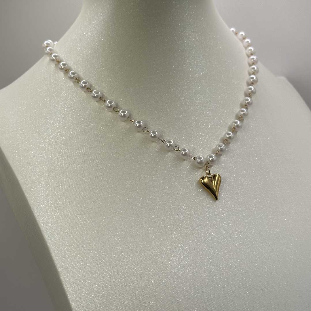 Ce magnifique bijou "Jane" en acier inoxydable est un collier de parles blanches avec un pendentif coeur de couleur or. Il est posé sur un buste pailleté de couleur blanc qui est lui même sur un fond blanc.