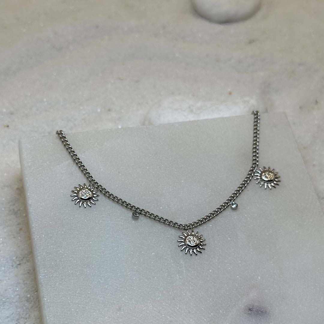 Un très beau collier en argent "june" est au centre de l'image, joliment posé sur une plaque de marbre. Le bijoux en acier est composé d'une chaîne en argent de 5 charms : 3 soleils et 2 strass. Autour se trouve du sable blanc.