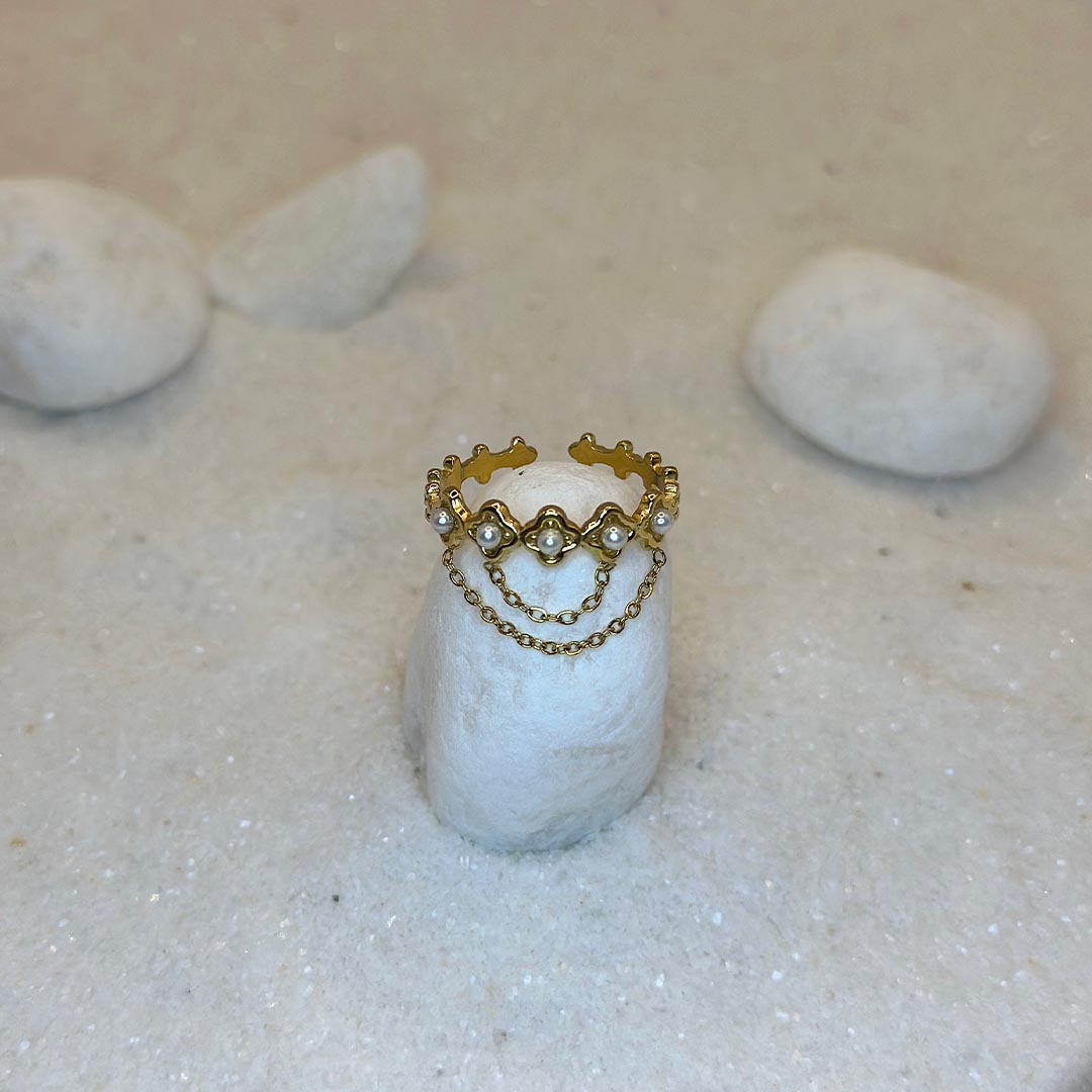 Une très belle bague en or "livia" est posée autour d'un caillou blanc, au centre de l'image. Le bijoux en acier inoxydable est aussi composé de 2 chaînes en or, reliées à des perles. Tout autour se trouve du sable blanc.
