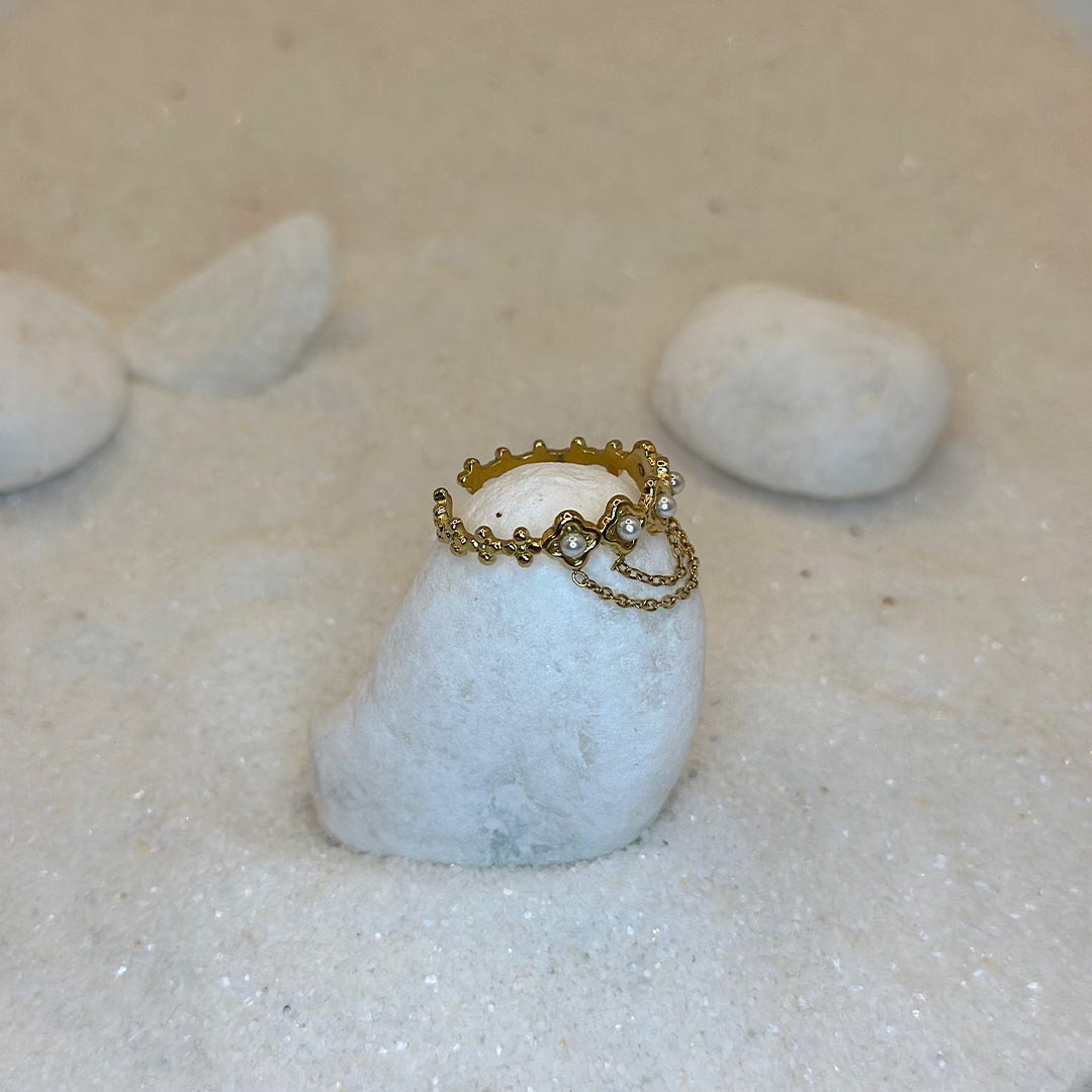 Une très belle bague en or "livia" est posée autour d'un caillou blanc, au cnetre de l'image vu de côté. Le bijoux en acier inoxydable est aussi composé de 2 chaînes en or, reliées à des perles. Tout autour se trouve du sable blanc.