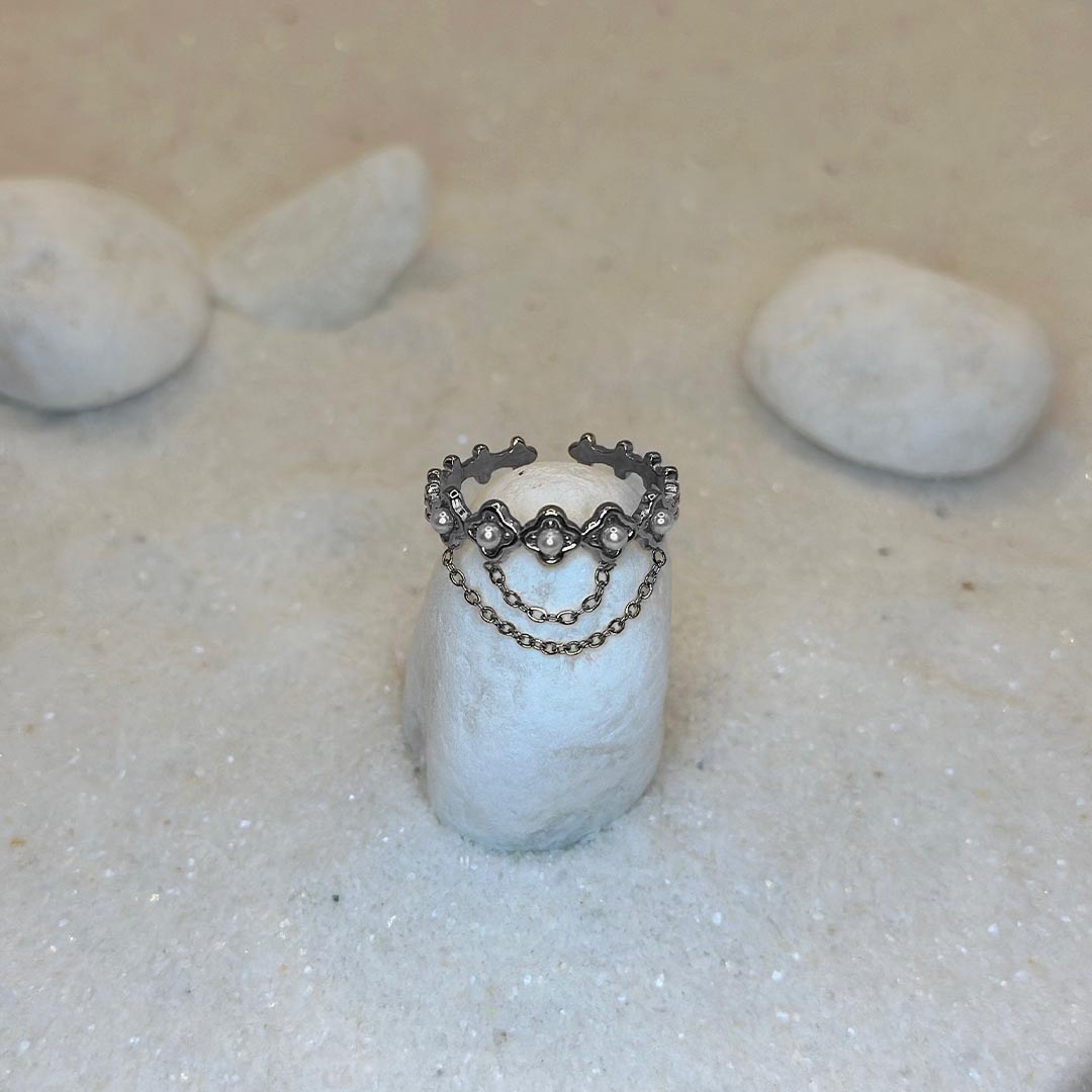 Une très belle bague argenté "livia" est posée autour d'un caillou blanc, au centre de l'image. Le bijoux en acier inoxydable est aussi composé de 2 chaînes argentées, reliées à des trèfles. Tout autour se trouve du sable blanc.
