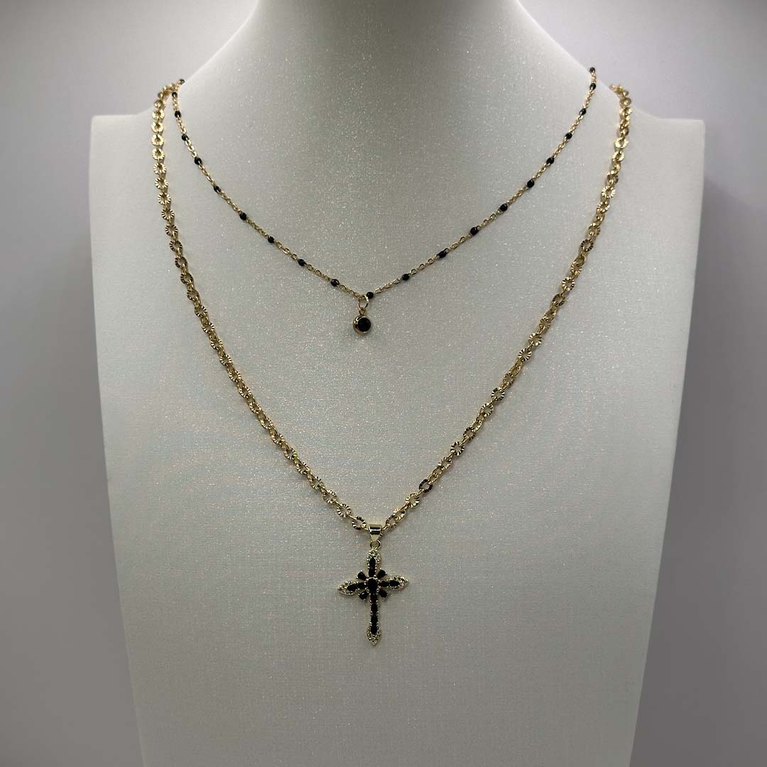 Ces jolis bijoux "Lola" et "crecia" en acier inoxydable sont des colliers avec une chaîne dorée et boules noires avec un petit pendentif noir à strass et l'autre avec une chaîne dorée et un pendentif croix noir a strass noirs. Ils sont posés sur un buste blanc pailleté qui est lui même posé devant un fond blanc.