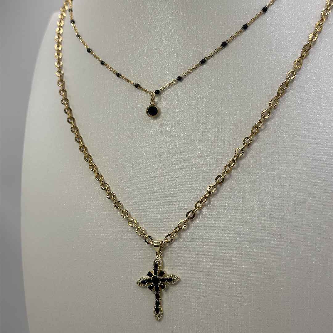 Ces jolis bijoux "Lola" et "crecia" en acier inoxydable sont des colliers avec une chaîne dorée et boules noires avec un petit pendentif noir à strass et l'autre avec une chaîne dorée et un pendentif croix noir a strass noirs. Ils sont posés sur un buste blanc pailleté qui est lui même posé devant un fond blanc.