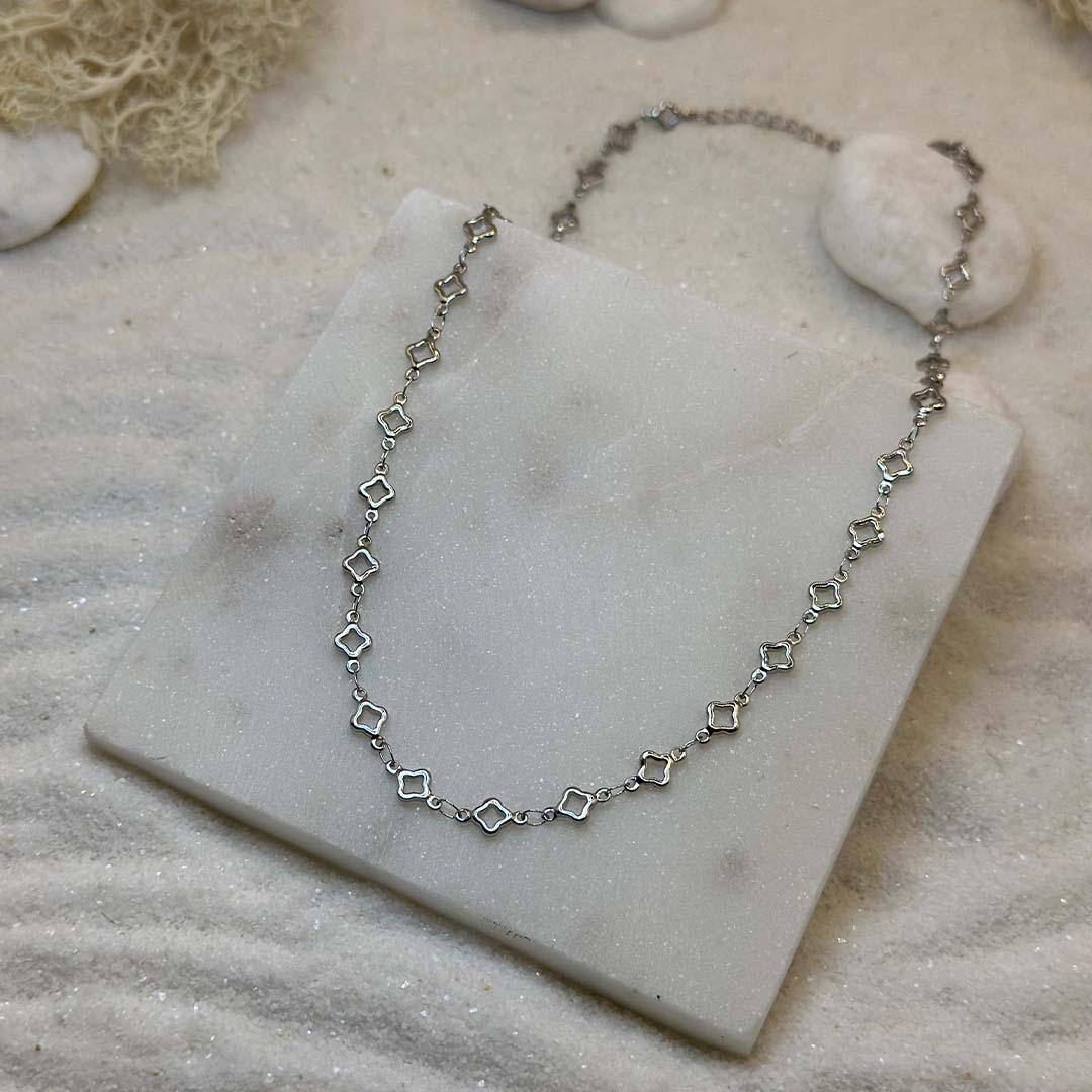 Un magnifique collier en argent "lori" est au centre de l'image, posé sur une plaque de marbre. Le bijoux en acier inoxydable est composé d'une chaîne en argent, ainsi que des motifs en forme d'étoiles. Autour se trouve du sable blanc.