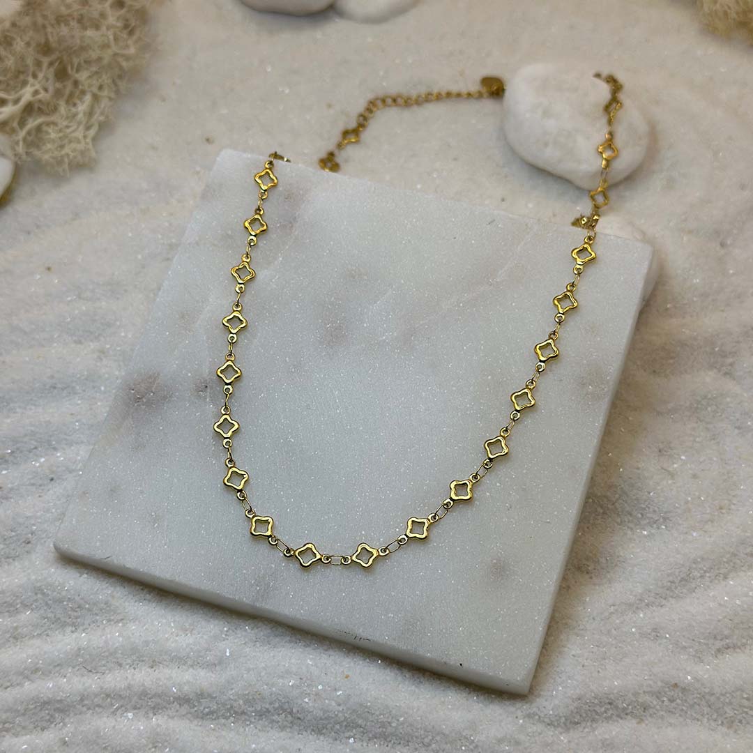 Un magnifique collier en or "lori" est au centre de l'image, posé sur une plaque de marbre. Le bijoux en acier inoxydable est composé d'une chaîne en or, ainsi que des motifs en forme d'étoiles. Autour se trouve du sable blanc.