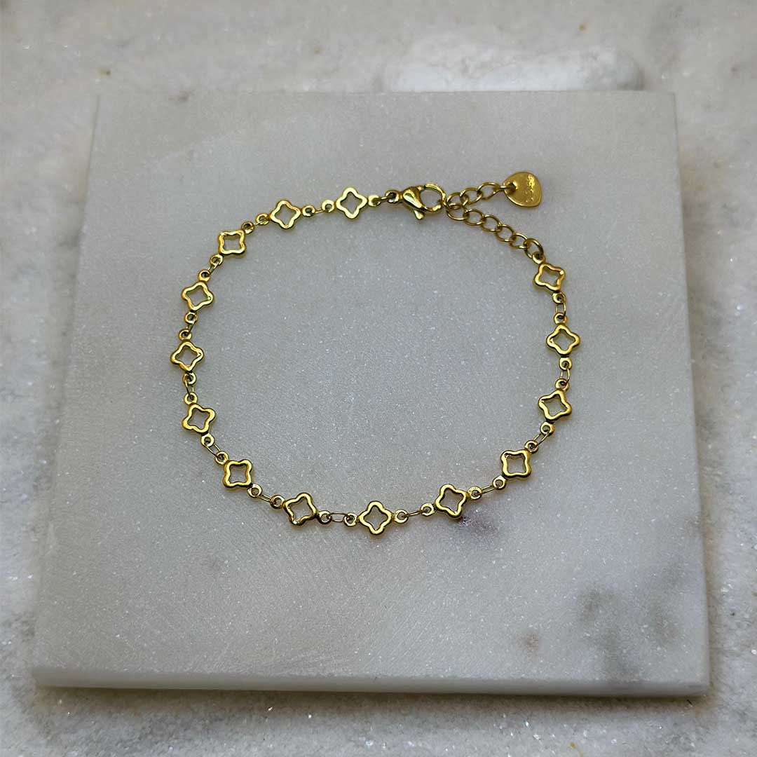 Un magnifique bracelet en or "lori" est au centre de l'image en gros plan, posé sur une plaque de marbre. Le bijoux en acier inoxydable est composé d'une chaîne en or, ainsi que des motifs en forme d'étoiles. Autour se trouve du sable blanc.