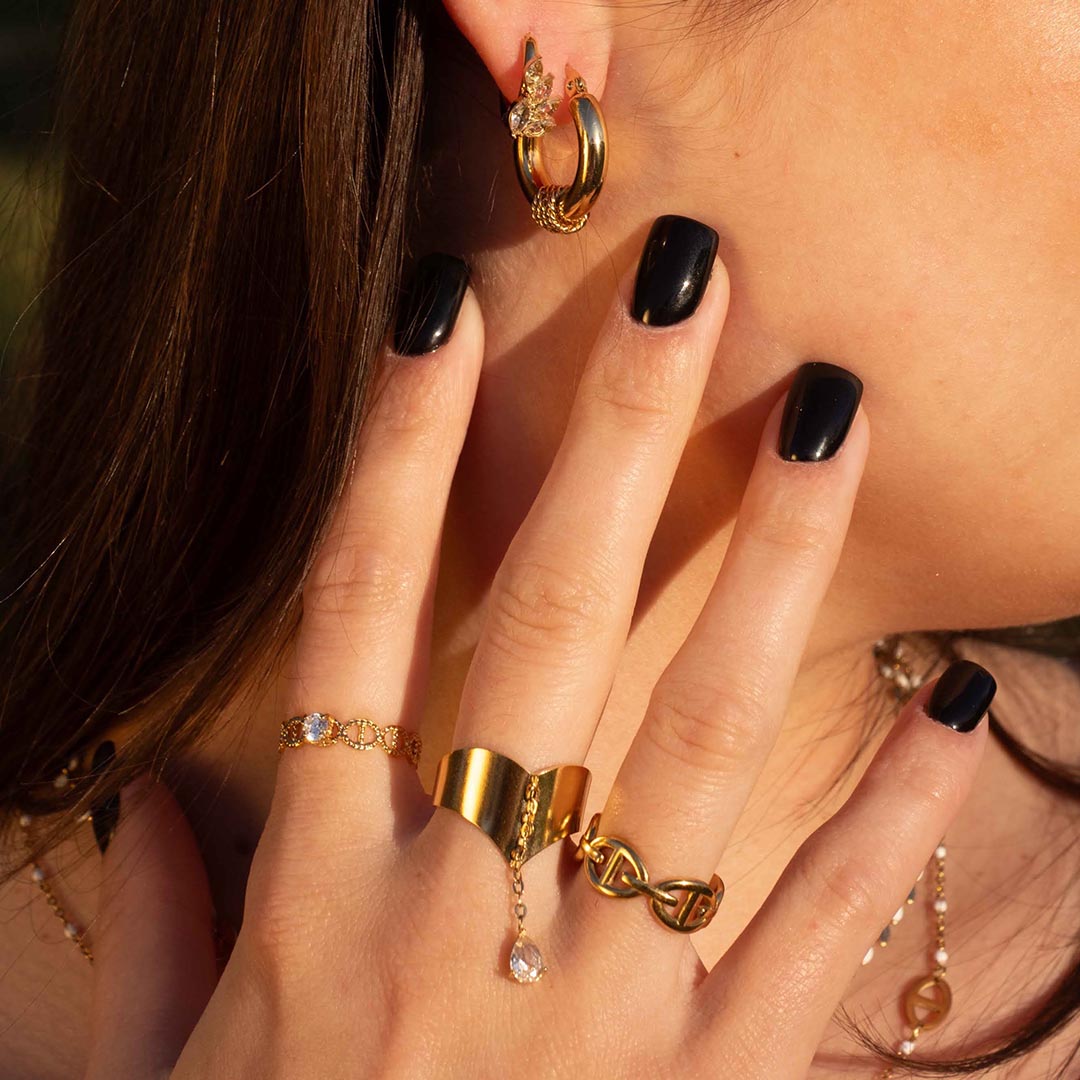 Photo d'une main montrant des bagues en acier inoxydable. On y retrouve la bague "ari" qui est avec un design de noeuds ou maillons marins, La bague "mira" au design de noeuds marins et strass. La bague "enea" avec une bague large reliant un strass par une chaînette. Les bijoux sont tous de couleur or / dorés. En arrière plan nous pouvons retrouver les cheveux bruns de la modèle qui porte les bijoux. Nous y voyons aussi les boucles d'oreilles "Lou" et "Ania" La lumière de la photo est chaude.