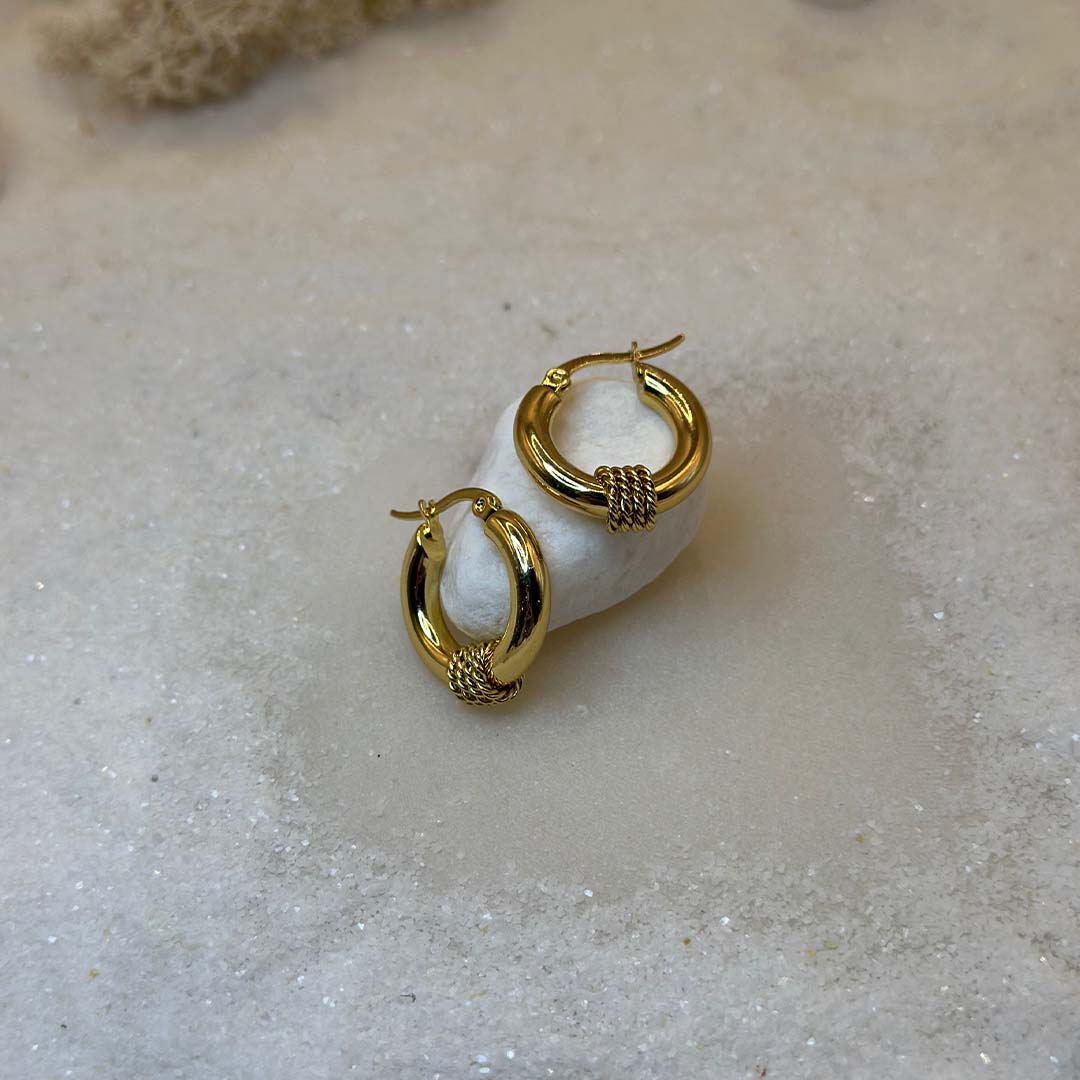 Une magnifique paire de boucles d'oreille en or "lou" est au centre de l'image, posée sur un cailloux blanc. Les bijoux en acier inoxydable sont vu de haut, composés d'une base lisse et de 3 cordes en relief au centre. Autour se trouve du sable blanc.