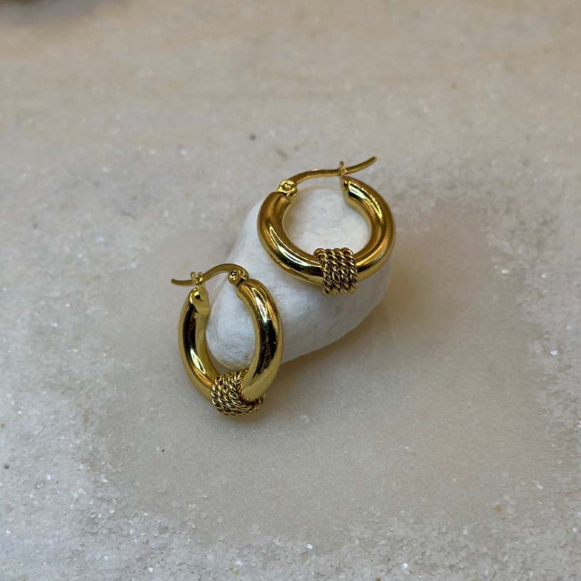 Une magnifique paire de boucles d'oreille en or "lou" est au centre de l'image, posée sur un cailloux blanc. Les bijoux en acier inoxydable sont vu de face, composés d'une base lisse et de 3 cordes en relief au centre. Autour se trouve du sable blanc.