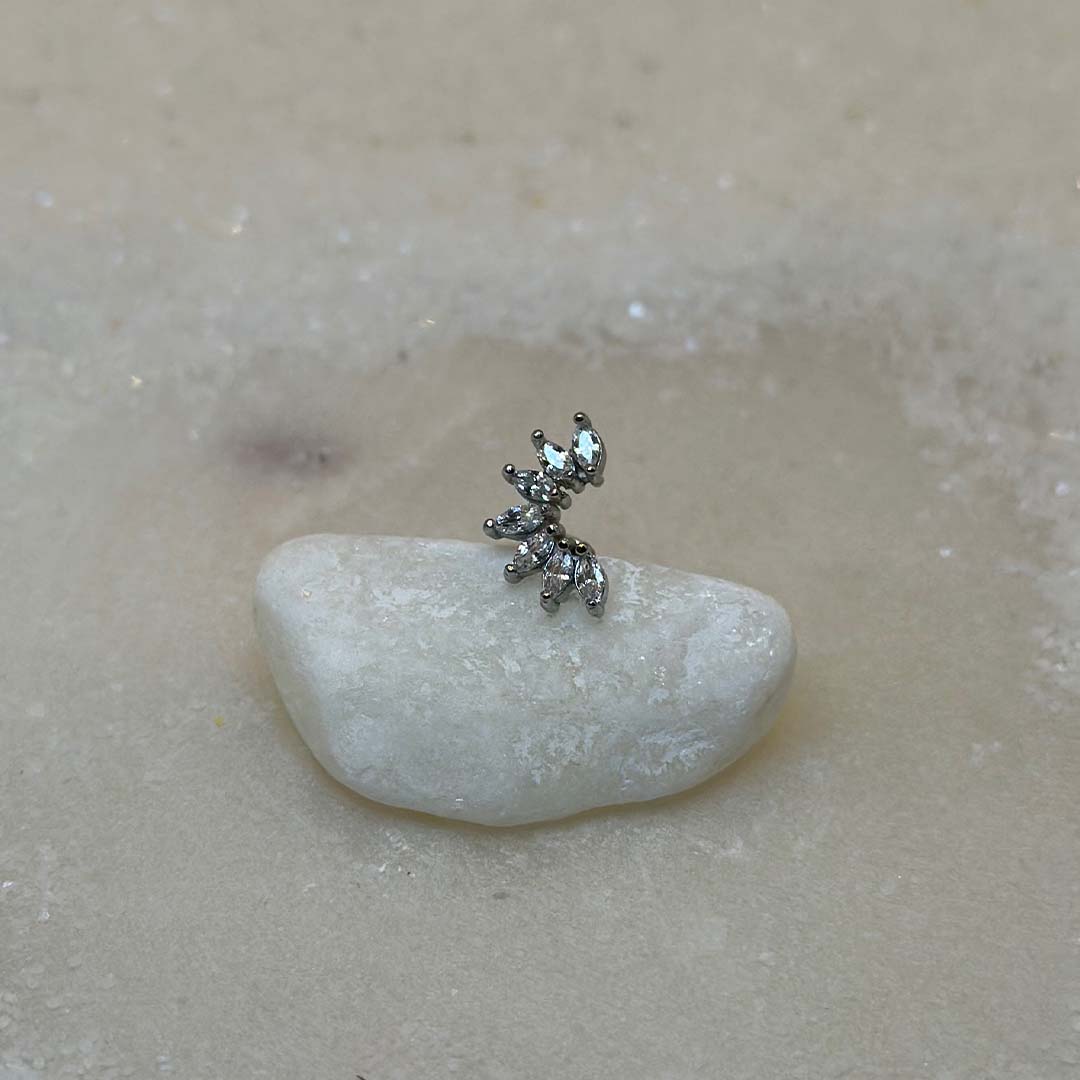 Un très beau piercing "Luisa", de couleur argent, est au centre de l'image. Le bijoux en acier inoxydable est posé de 3/4 sur un caillou blanc. Le fond est du sable blanc.