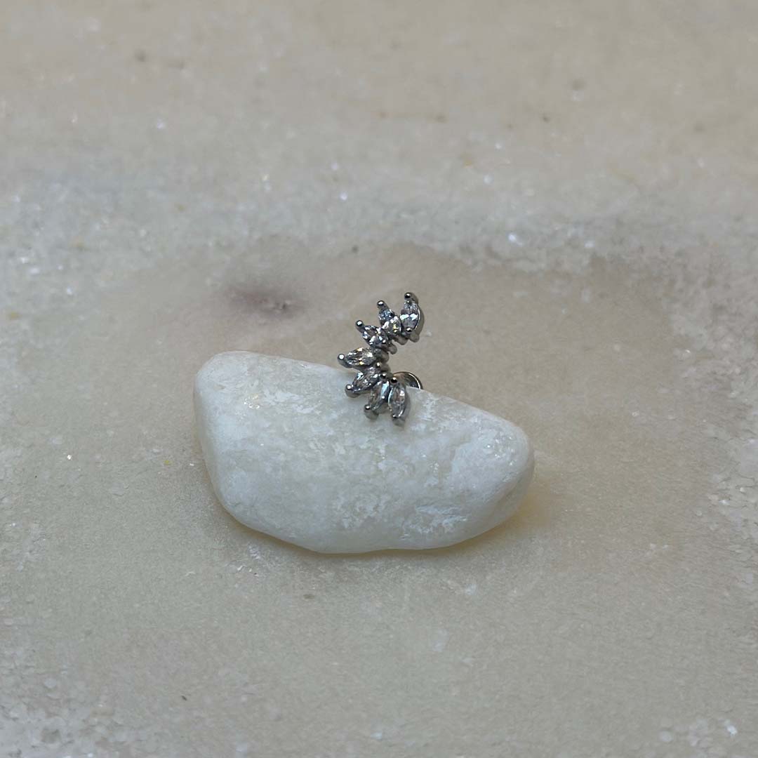 Un très beau piercing "Luisa", de couleur argent, est au centre de l'image. Le bijoux en acier inoxydable est posé de 3/4 sur un caillou blanc. Le fond est du sable blanc.