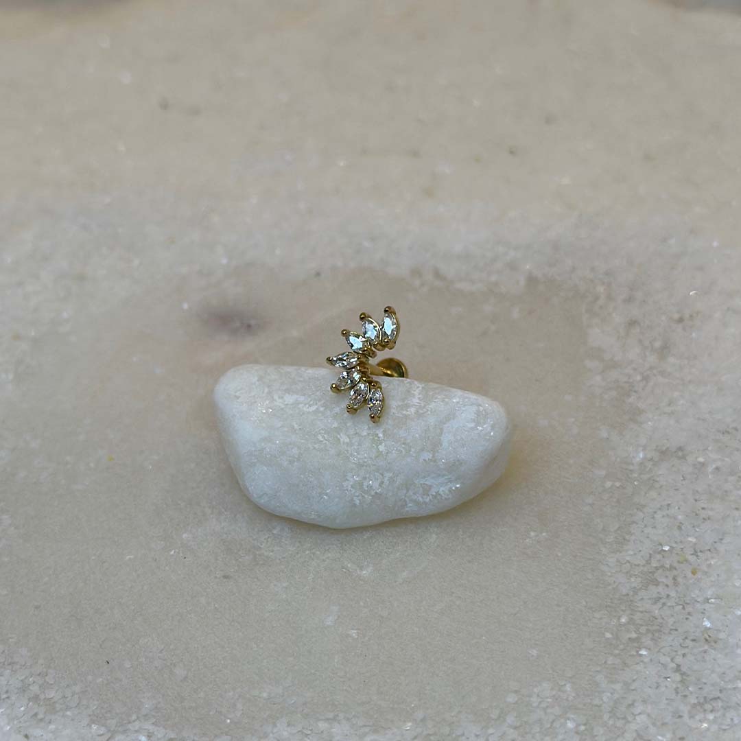 Un très beau piercing "Luisa", de couleur or, est au centre de l'image. Le bijoux en acier inoxydable est posé de 3/4 sur un caillou blanc. Le fond est du sable blanc.
