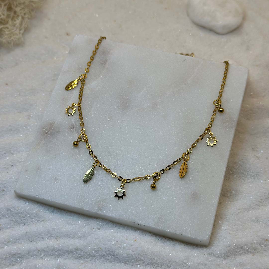 Un magnifique collier en acier inoxydable "Lumina" de couleur or, ce bijou possède plusieurs charms : plumes, soleils ainsi que des perles couleur or, ce bijou est posé sur une plaque de marbre blanche qui elle même est posé sur du sable blanc qui fait office de fond d'image.