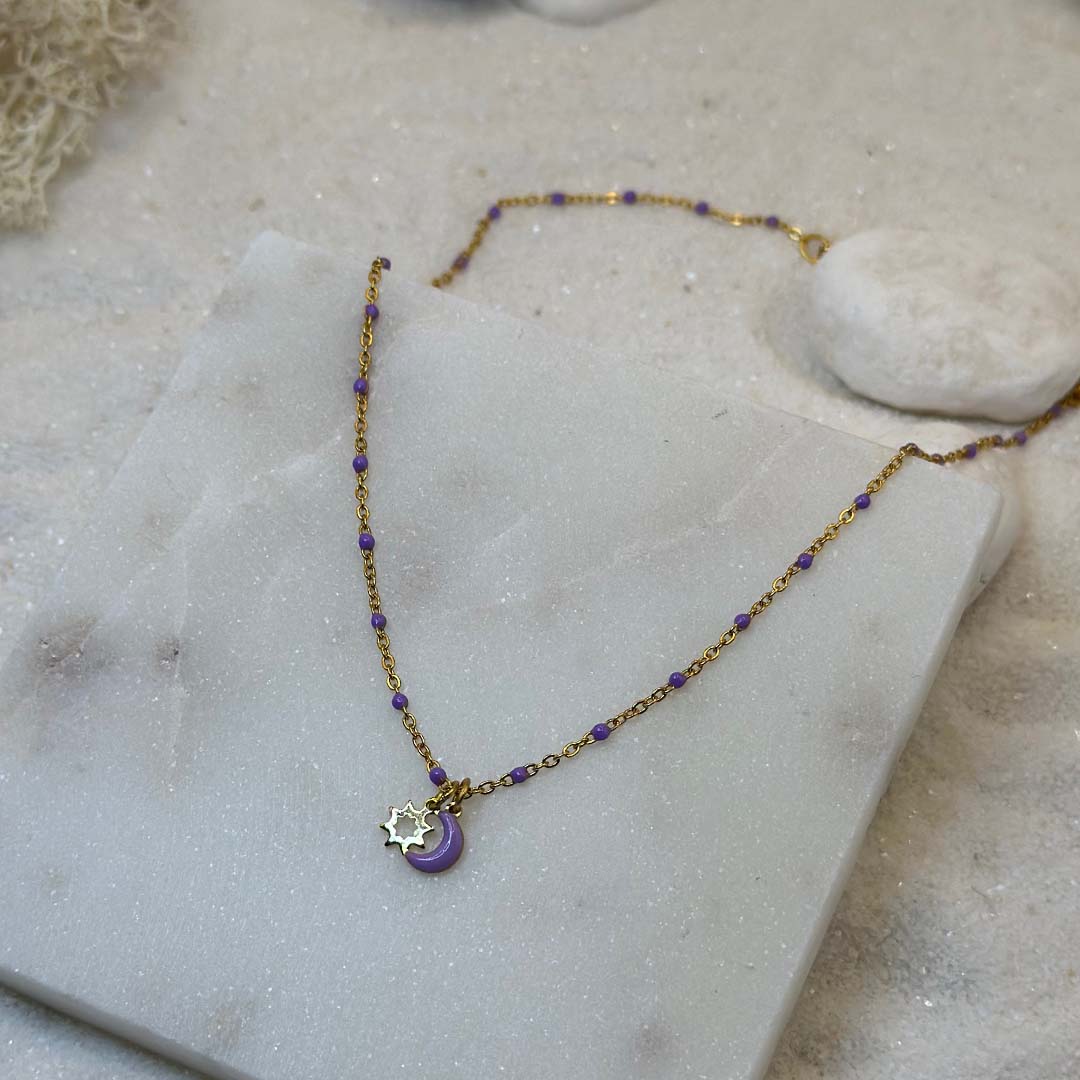 Un magnifique collier en acier inoxydable "Lunicia" de couleur or et violet, ce bijou possède pendentifs lune et soleils de couleurs or et violet ainsi que plusieurs perles violettes tout le long de la chaine, ce bijou est posé sur une plaque de marbre blanche qui elle même est posé sur du sable blanc qui fait office de fond d'image.