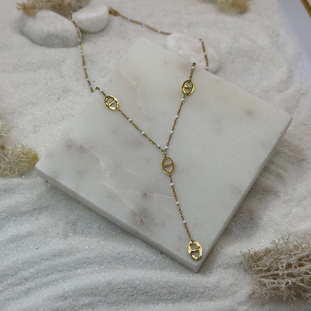 Un magnifique collier en acier inoxydable "Mada" de couleur or et et blanc, ce bijou possède plusieurs pendentifs de forme noeuds marin et des perles blanches tout le long de la chaine, ce bijou est posé sur une plaque de marbre blanche qui elle même est posé sur du sable blanc qui fait office de fond d'image.