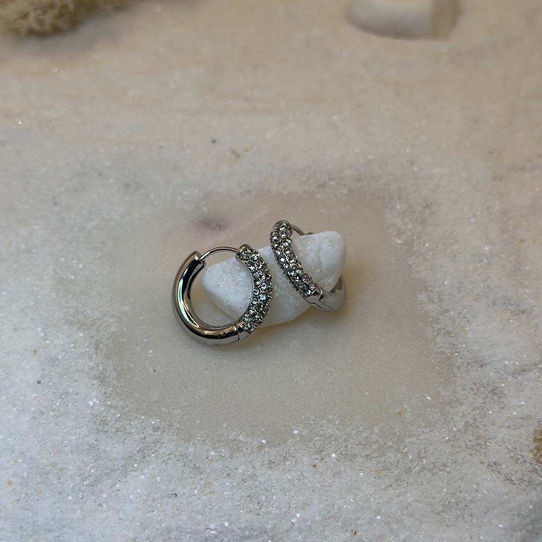 Une magnifique paire de boucle d'oreilles en acier inoxydable "Mandy" de couleur argent, ces boucles d'oreilles sont de forme ronde avec un coté en strass tte bagues scindé en deux, ce bijou est posé sur un caillou blanc qui est posé sur du sable blanc qui fait office de fond d'image.