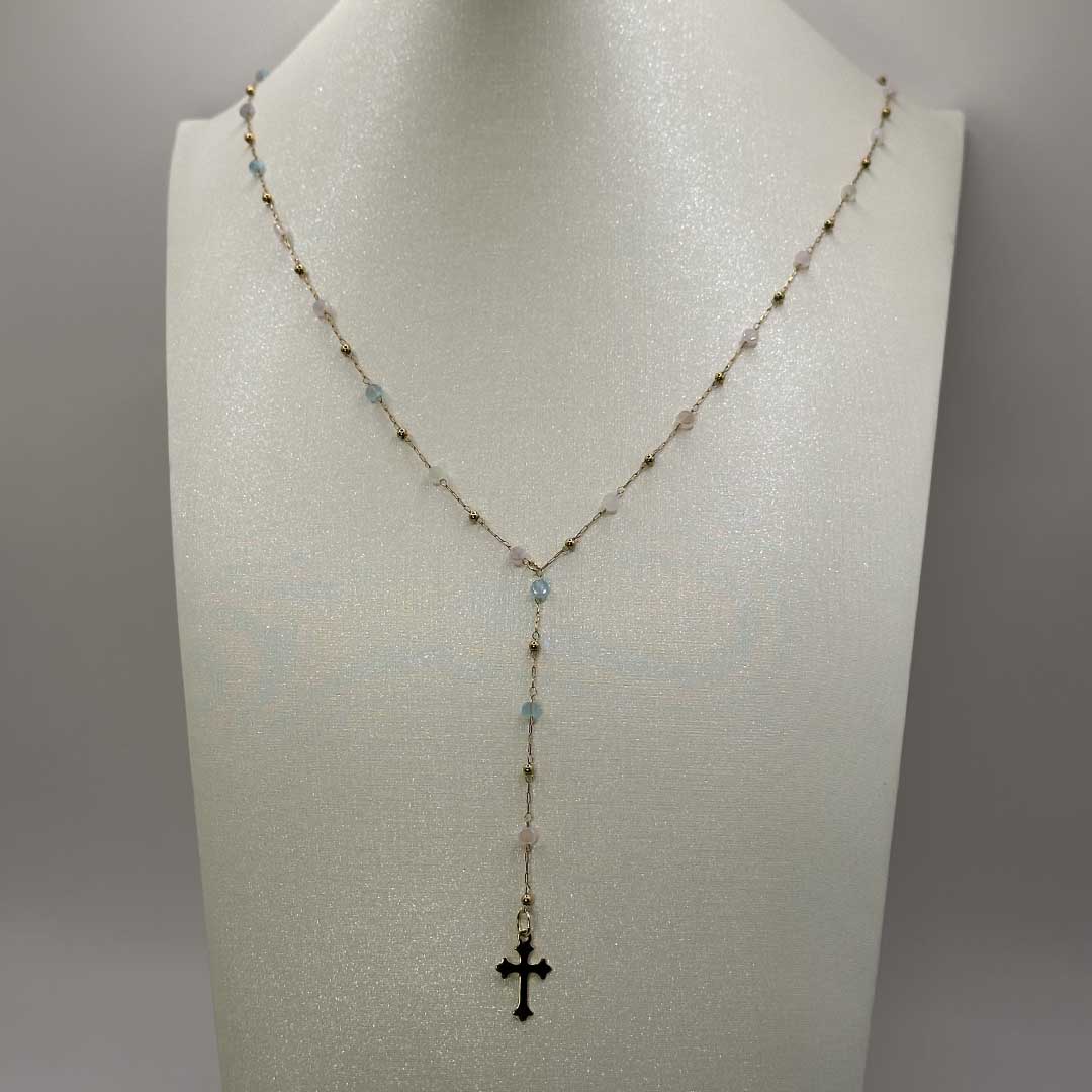 Ce joli bijou "mia" en acier inoxydable est un collier Y ou lasso avec une chaîne dorée et des pierres colorées en bleu, rose et vert. Il y a des petites boules de couleur or sur la chaîne et au bout se trouve en pendentif croix doré style gothique. Le collier est sur un buste pailleté blanc qui est devant un fond blanc.