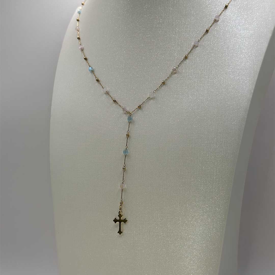 Ce joli bijou "mia" en acier inoxydable est un collier Y ou lasso avec une chaîne dorée et des pierres colorées en bleu, rose et vert. Il y a des petites boules de couleur or sur la chaîne et au bout se trouve en pendentif croix doré style gothique. Le collier est sur un buste pailleté blanc qui est devant un fond blanc.
