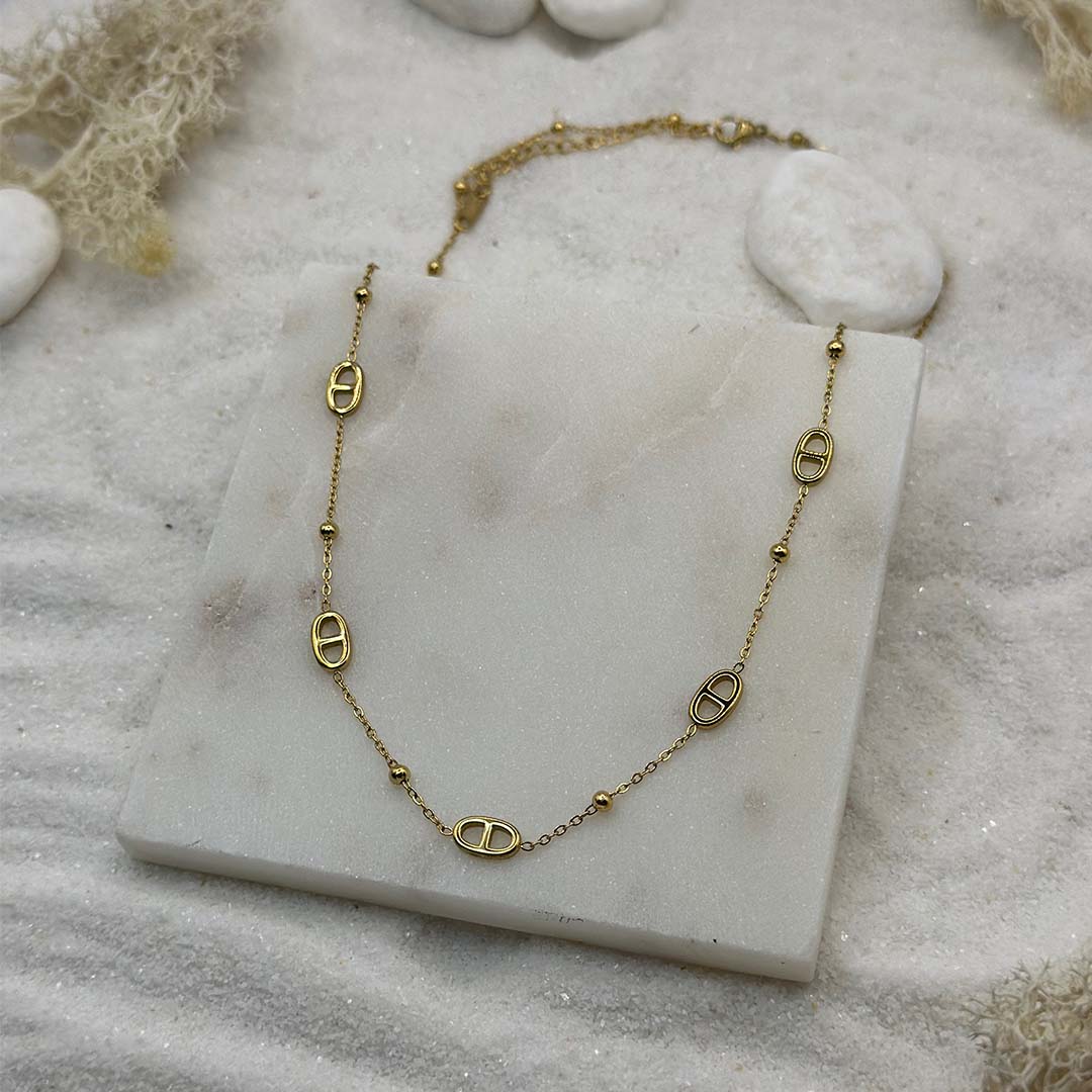 Un magnifique collier en acier inoxydable "Mina" de couleur or, ce bijou possède plusieurs nouer marin ainsi que des perles couleur or, ce bijou est posé sur une plaque de marbre blanche qui elle même est posé sur du sable blanc qui fait office de fond d'image.