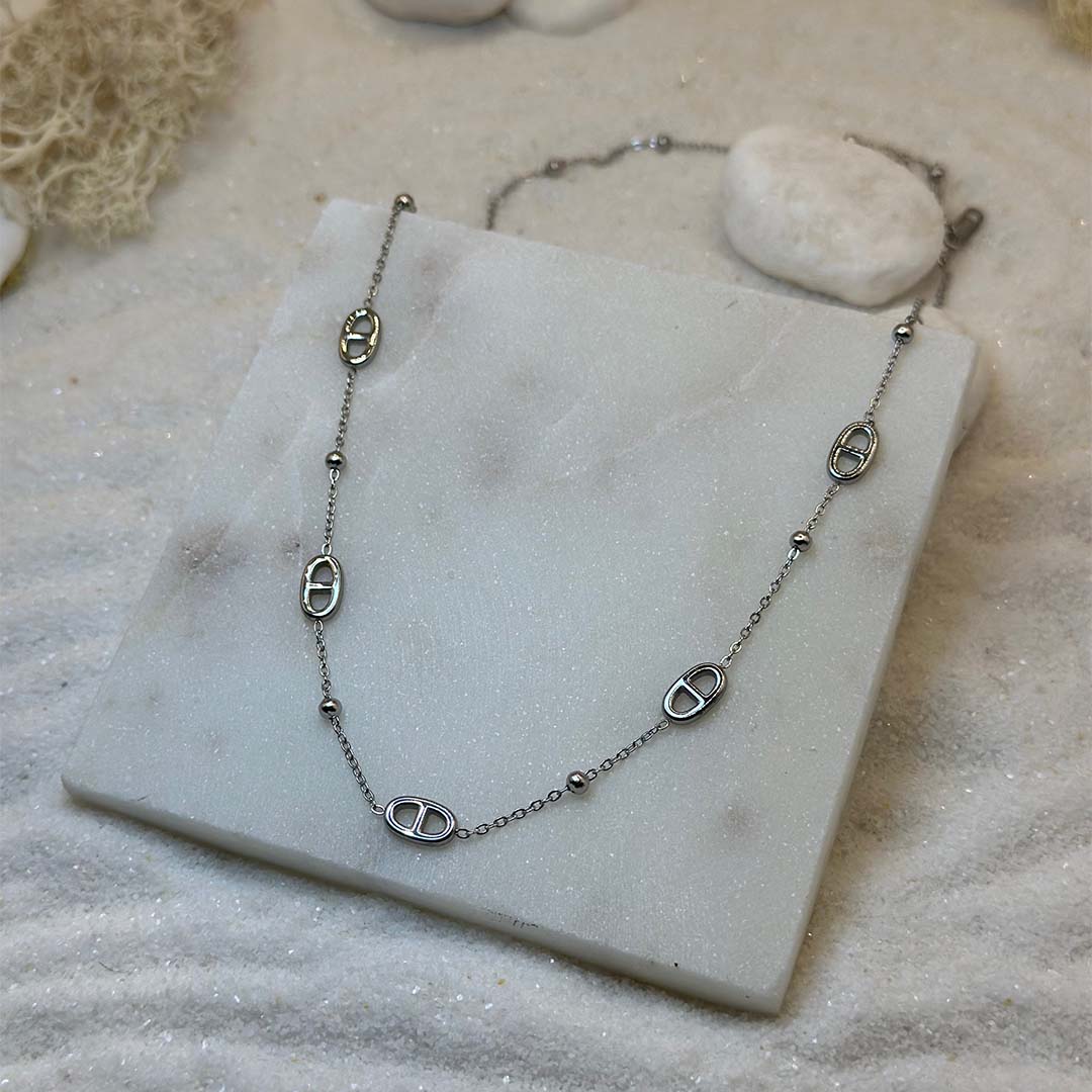 Un magnifique collier en acier inoxydable "Mina" de couleur argent, ce bijou possède plusieurs nouer marin ainsi que des perles couleur argent, ce bijou est posé sur une plaque de marbre blanche qui elle même est posé sur du sable blanc qui fait office de fond d'image.