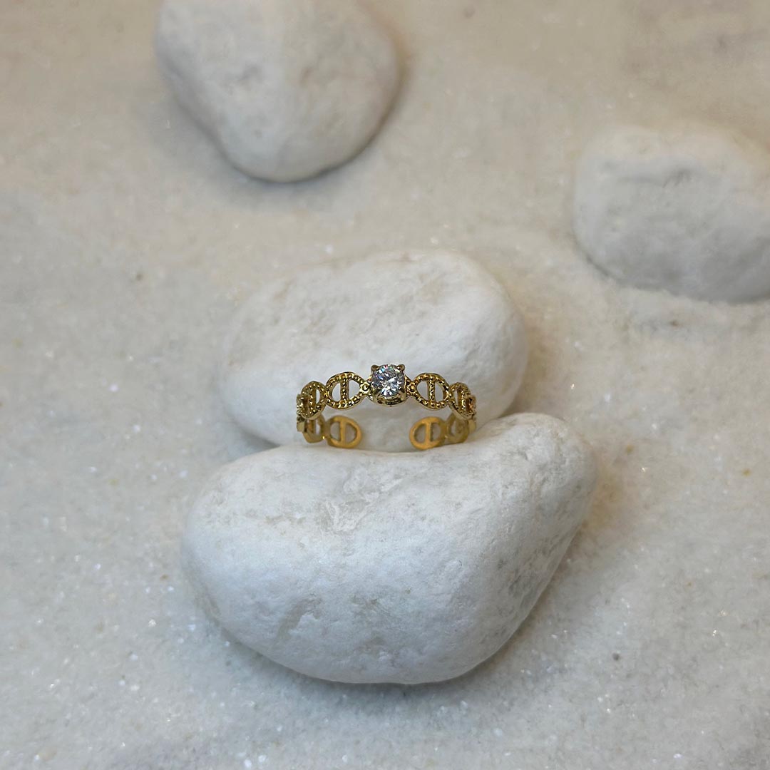 Une magnifique bague en acier inoxydable "Mira" de couleur or, cette bague est de forme ronde avec des noeud marin sur tout le tour et avec un strass au centre, le bijou est posé sur un caillou blanc qui est posé sur du sable blanc qui fait office de fond d'image.