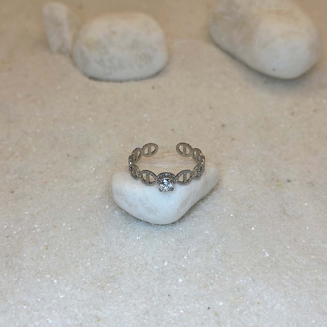 Une magnifique bague en acier inoxydable "Mira" de couleur argent, cette bague est de forme ronde avec des noeud marin sur tout le tour et avec un strass au centre, le bijou est posé sur un caillou blanc qui est posé sur du sable blanc qui fait office de fond d'image.