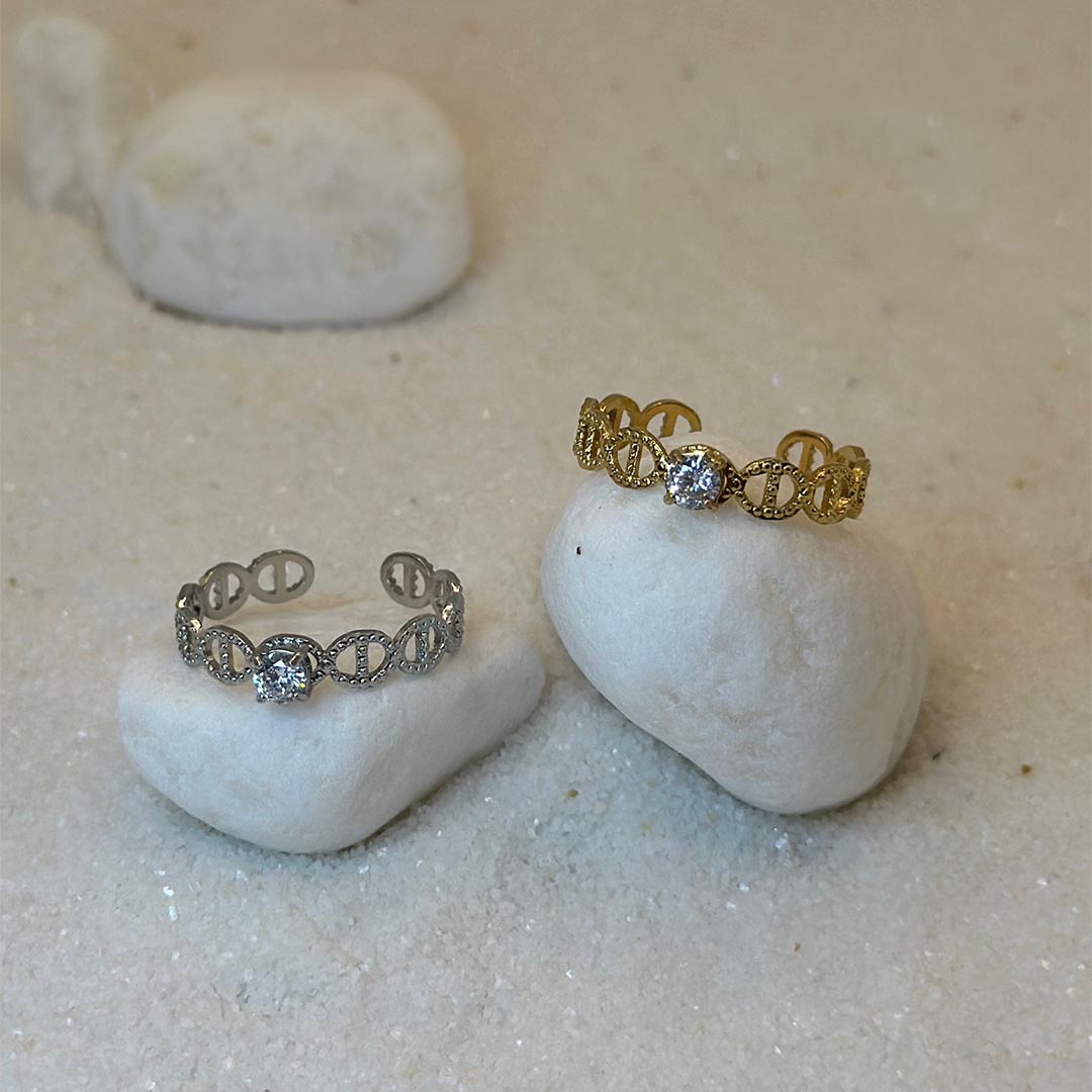 Deux magnifique bagues en acier inoxydable "Mira" de couleur argent et or, ces bagues sont de forme ronde avec deux noeud marin sur tout leurs tours et avec un strass en leurs centre, ces bijoux sont posé sur un caillou blanc qui est posé sur du sable blanc qui fait office de fond d'image.