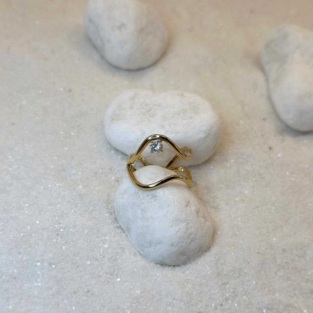 Un magnifique bague en acier inoxydable "Noeline" de couleur or, cette bagues scindé en deux, elle est de forme irrégulière avec un strass entre les deux formes, ce bijou est posé sur un caillou blanc qui est posé sur du sable blanc qui fait office de fond d'image.