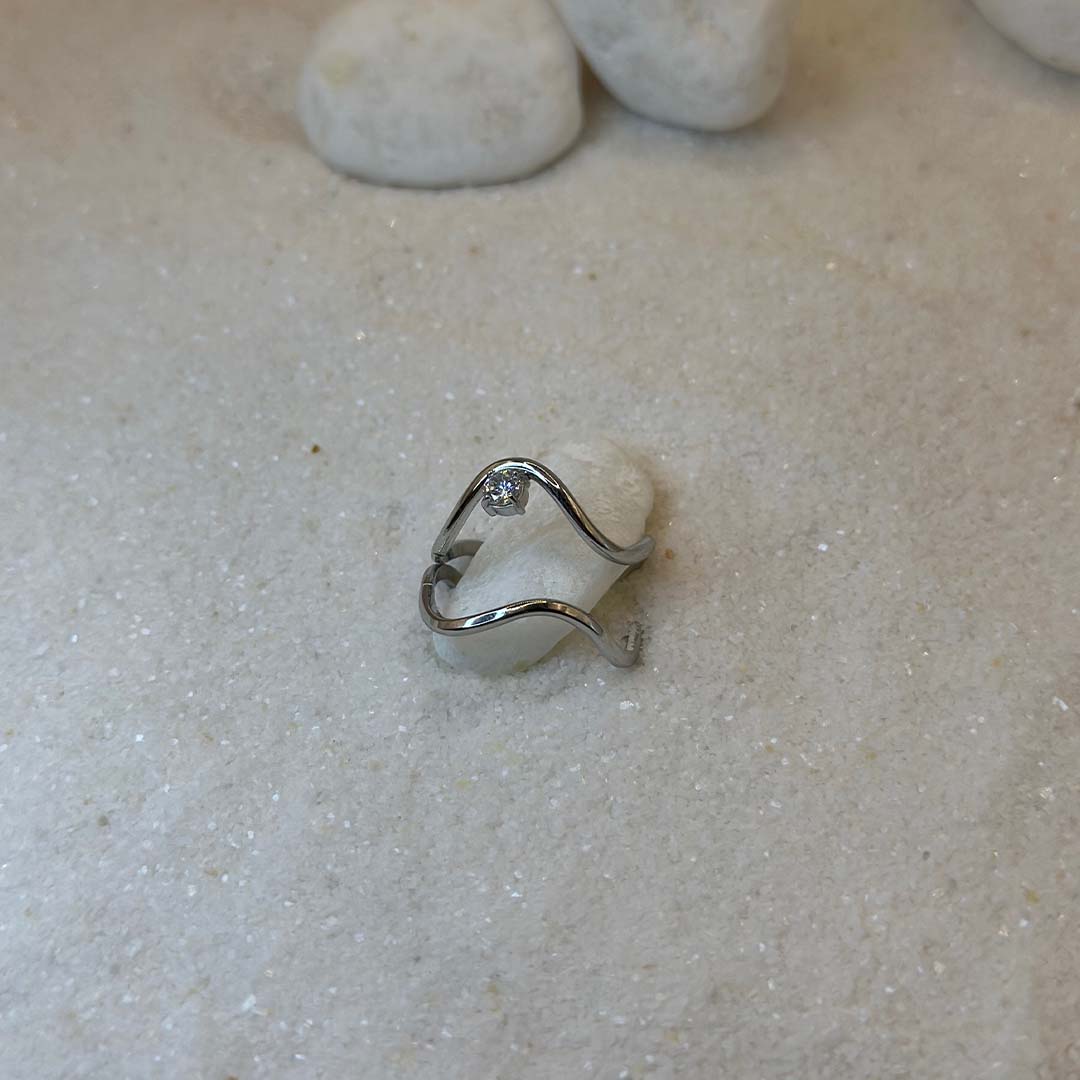 Un magnifique bague en acier inoxydable "Olivia" de couleur argent, cette bagues scindé en deux, elle est de forme irrégulière avec un strass entre les deux formes, ce bijou est posé sur un caillou blanc qui est posé sur du sable blanc qui fait office de fond d'image.