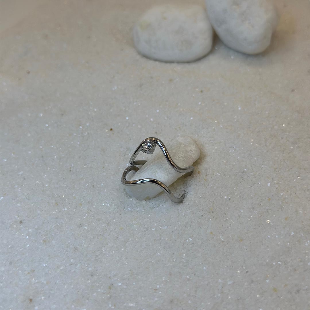 Un magnifique bague en acier inoxydable "Noeline" de couleur argent, cette bagues scindé en deux, elle est de forme irrégulière avec un strass entre les deux formes, ce bijou est posé sur un caillou blanc qui est posé sur du sable blanc qui fait office de fond d'image.