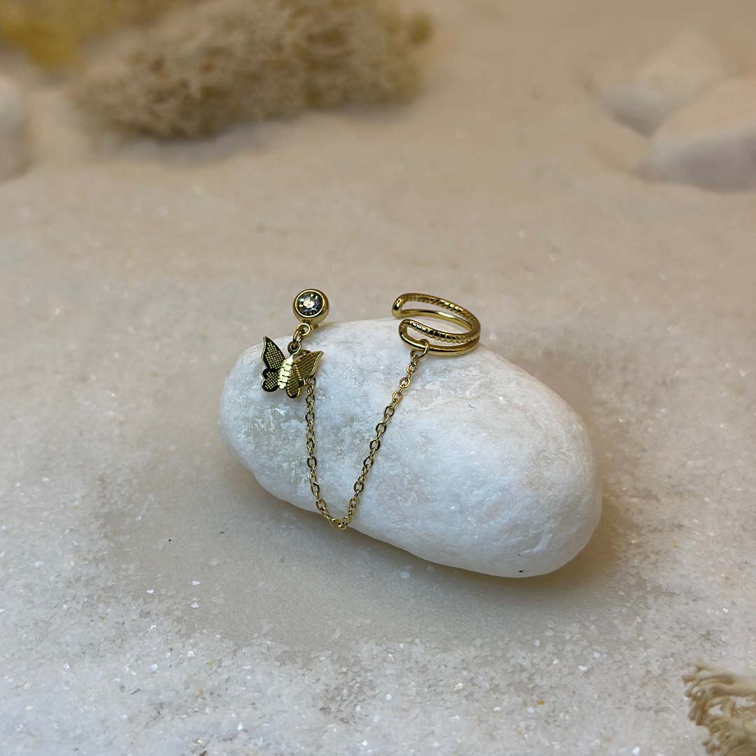 Un magnifique paire de boucle d'oreilles en acier inoxydable "Palma" de couleur Or, le bijou possède d'un cote un strass avec un papillon en pendentif ainsi qu'une bague de l'autre et est relié par une chaine, ce bijou est posé sur un caillou blanc qui est posé sur du sable blanc qui fait office de fond d'image.