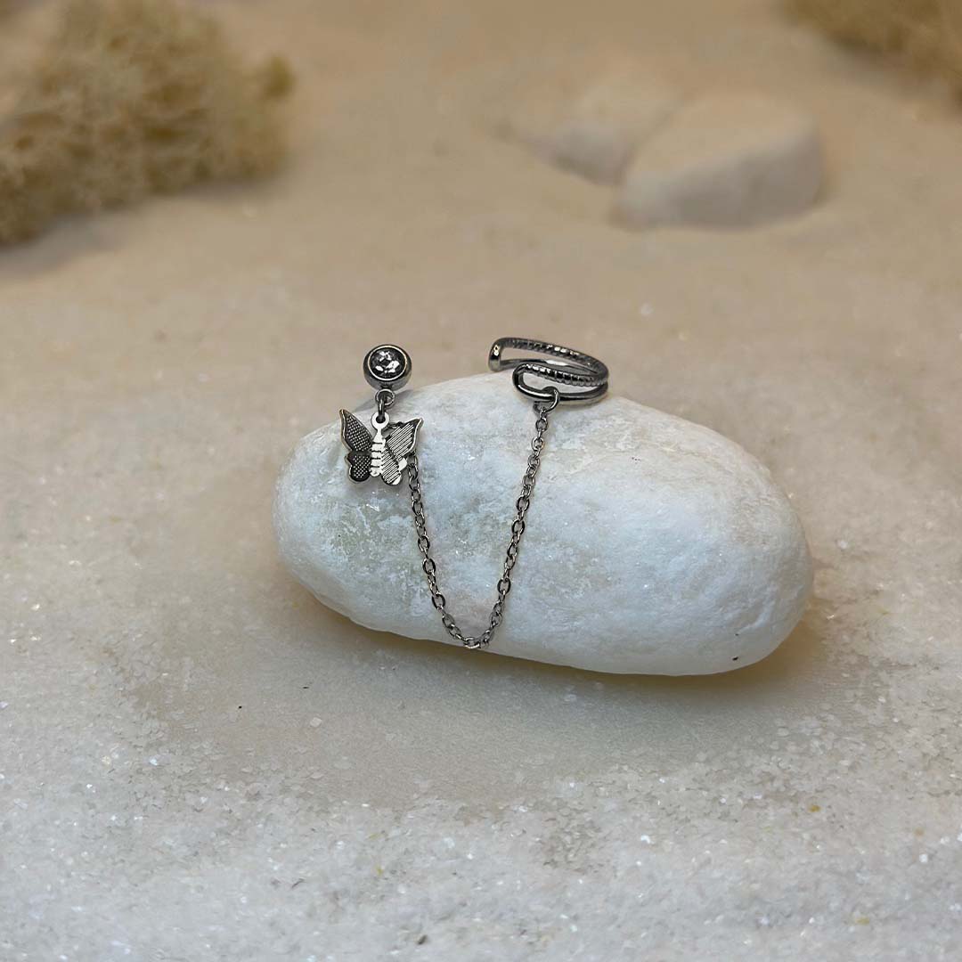 Un magnifique paire de boucle d'oreilles en acier inoxydable "Palma" de couleur Argenté, le bijou possède d'un cote un strass avec un papillon en pendentif ainsi qu'une bague de l'autre et est relié par une chaine, ce bijou est posé sur un caillou blanc qui est posé sur du sable blanc qui fait office de fond d'image.