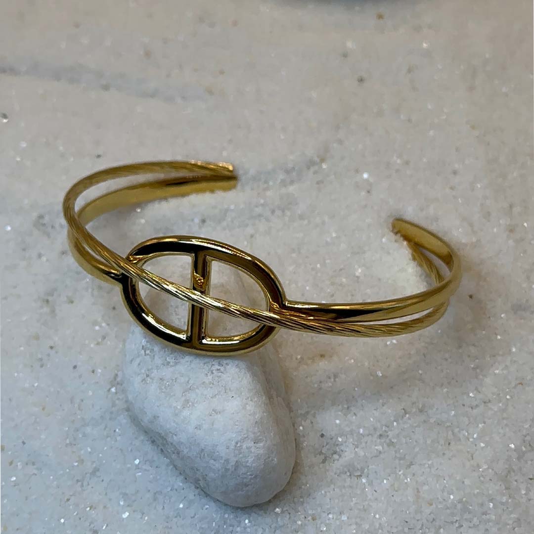 Un magnifique Jonc en acier inoxydable "Romy" de couleur or, ce bijou possède une forme en noeud marin au centre, ce bijou est posé sur une plaque de marbre blanche qui elle même est posé sur du sable blanc qui fait office de fond d'image.