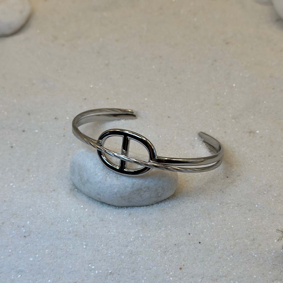 Un magnifique Jonc en acier inoxydable "Romy" de couleur argent, ce bijou possède une forme en noeud marin au centre, ce bijou est posé sur une plaque de marbre blanche qui elle même est posé sur du sable blanc qui fait office de fond d'image.