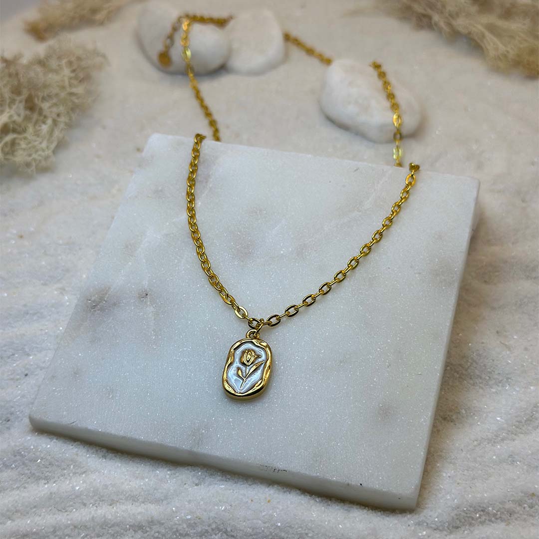 Un magnifique collier en acier inoxydable "Rosie" de couleur Or, le bijou possède un pendentif de forme ronde avec un intérieur blanc et une rose au centre, ce bijou est posé sur une plaque de marbre blanche qui elle même est posé sur du sable blanc qui fait office de fond d'image.