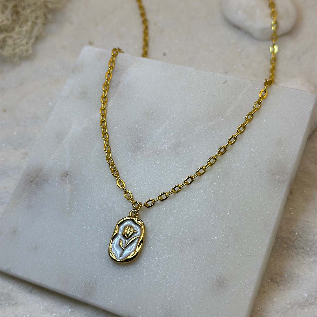 Un magnifique collier en acier inoxydable "Rosie" de couleur Or, le bijou possède un pendentif de forme ronde avec un intérieur blanc et une rose au centre, ce bijou est posé sur une plaque de marbre blanche qui elle même est posé sur du sable blanc qui fait office de fond d'image.