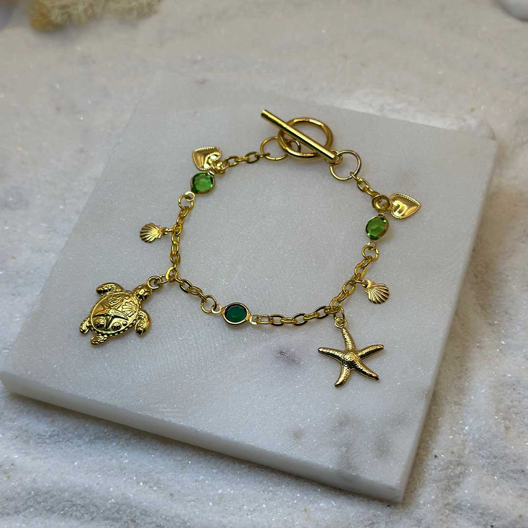 Un magnifique bracelet en acier inoxydable "Sania" de couleur or, ce bijou possède plusieurs charms : une étoile de mer, une tortue, un coquillage ainsi que deux coeurs, ci bijou est posé sur une plaque de marbre blanche qui elle même est posé sur du sable blanc qui fait office de fond d'image.