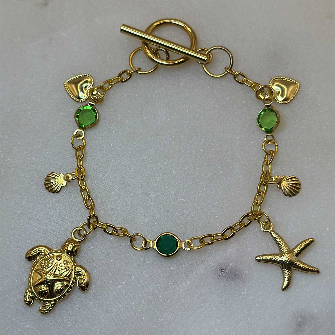 Un magnifique bracelet en acier inoxydable "Sania" de couleur or, ce bijou possède plusieurs charms : une étoile de mer, une tortue, un coquillage ainsi que deux coeurs, ci bijou est posé sur une plaque de marbre blanche qui elle même est posé sur du sable blanc qui fait office de fond d'image.