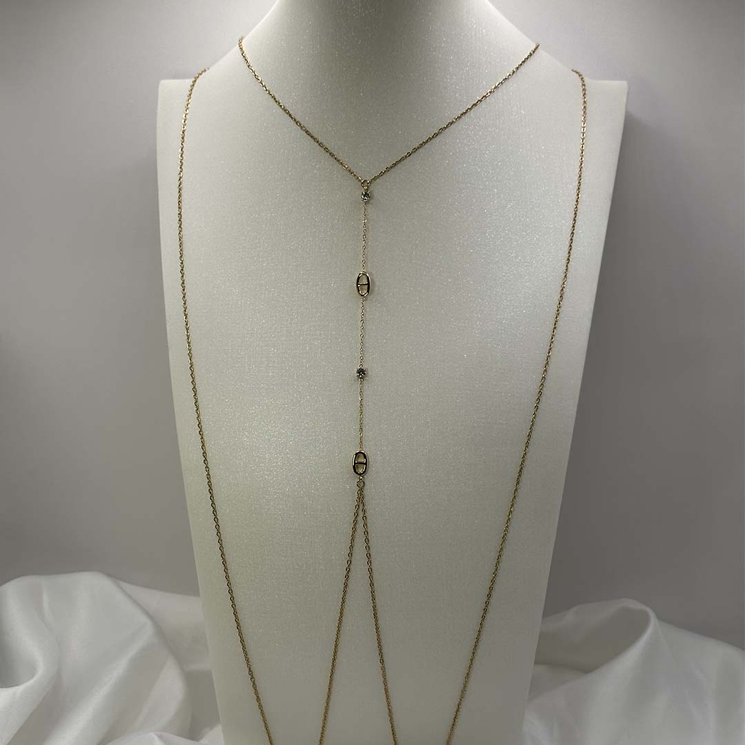 Ce magnifique bijou "Sélia" en acier inoxydable est un bijou de corps / une chaîne de buste dorée avec des strass et des charms au design de noeuds marins. Il est posé sur un buste blanc pailleté qui est sur une fond blanc et un tissu satiné blanc.