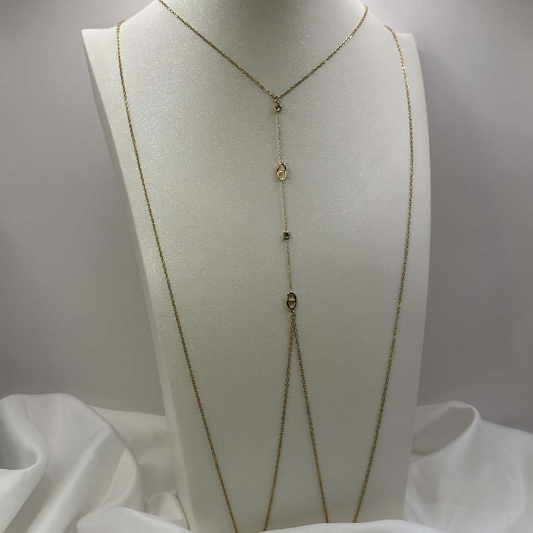 Ce magnifique bijou "Sélia" en acier inoxydable est un bijou de corps / une chaîne de buste dorée avec des strass et des charms au design de noeuds marins. Il est posé sur un buste blanc pailleté qui est sur une fond blanc et un tissu satiné blanc.