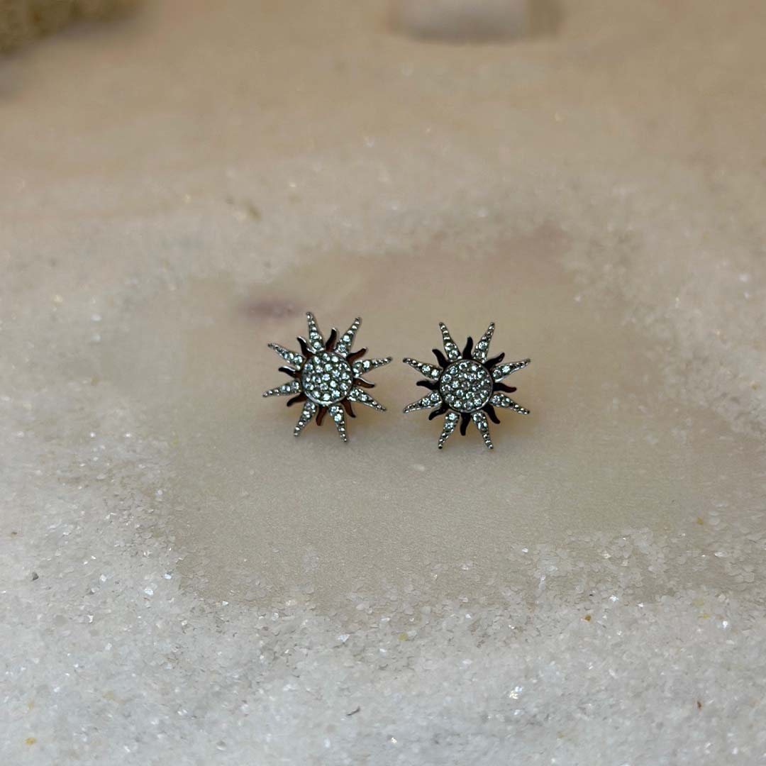 Un magnifique paire de boucle d'oreilles en acier inoxydable "Solea" de couleur Argent, le bijou possède une forme de soleil et est incrusté de diamants, ce bijou est posé sur une plaque de marbre blanche qui elle même est posé sur du sable blanc qui fait office de fond d'image.