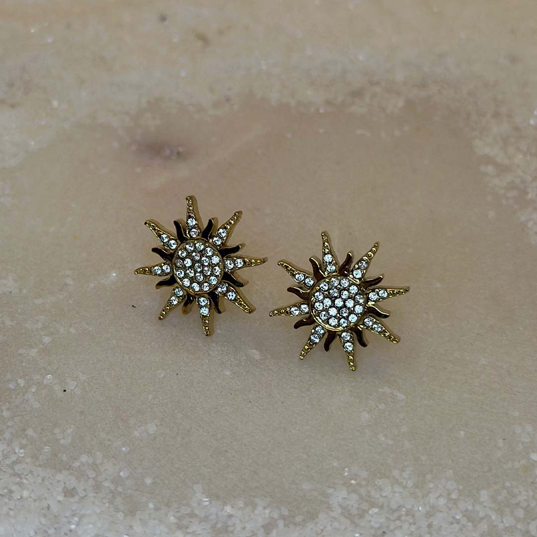 Un magnifique paire de boucle d'oreilles en acier inoxydable "Solea" de couleur Or, le bijou possède une forme de soleil et est incrusté de diamants, ce bijou est posé sur une plaque de marbre blanche qui elle même est posé sur du sable blanc qui fait office de fond d'image.