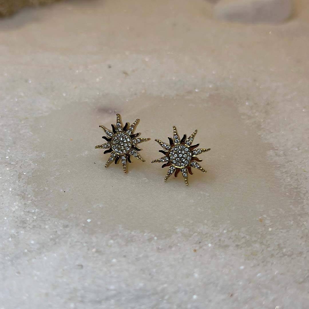 Un magnifique paire de boucle d'oreilles en acier inoxydable "Solea" de couleur Or, le bijou possède une forme de soleil et est incrusté de diamants, ce bijou est posé sur une plaque de marbre blanche qui elle même est posé sur du sable blanc qui fait office de fond d'image.